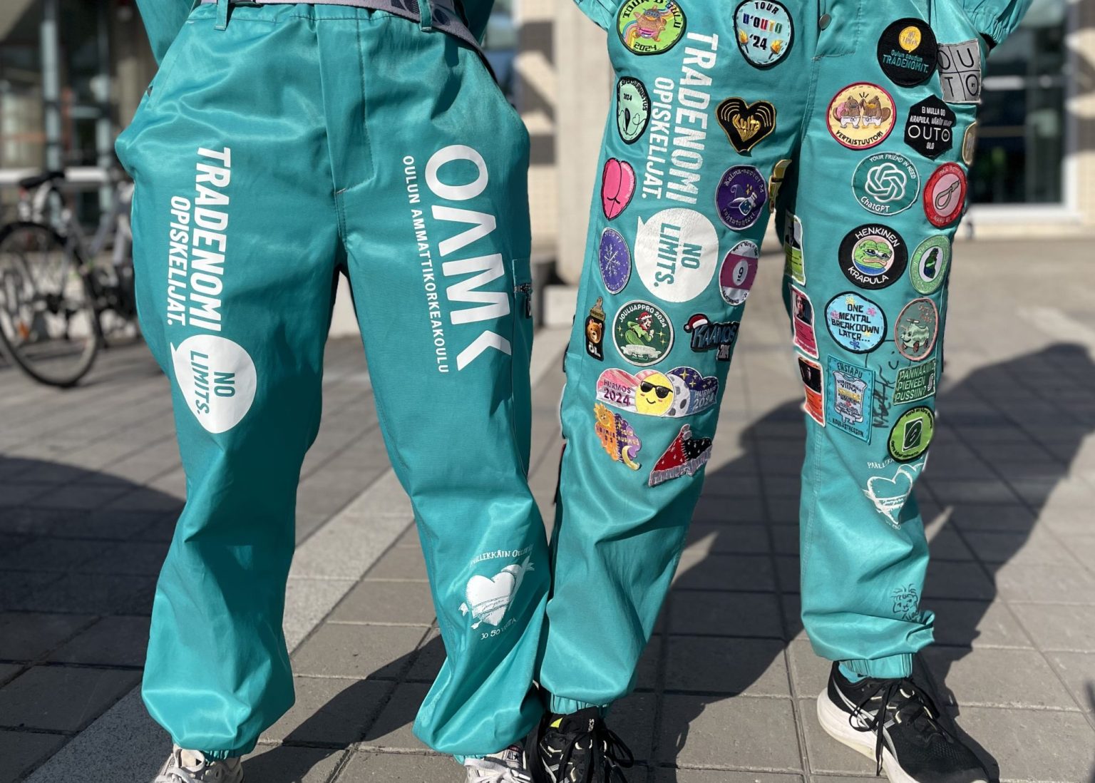 Kaksi henkilöä poseeraa turkoosit OUTO RY:n haalarit päällä ulkona. Toisissa haalareissa on haalarimerkkejä.