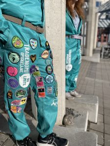 Kaksi henkilöä poseeraa turkoosit OUTO RY:n haalarit päällä ulkona. Toisissa haalareissa on haalarimerkkejä.