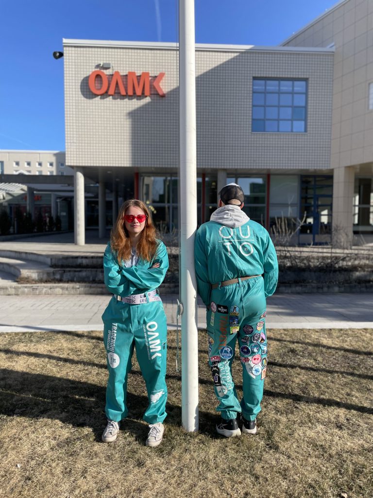 Kaksi henkilöä poseeraa turkoosit OUTO RY:n haalarit päällä ulkona. Toinen henkilöistä on kasvot päin kameraan, toinen henkilöistä on selin kameraan. Toisissa haalareissa on haalarimerkkejä.