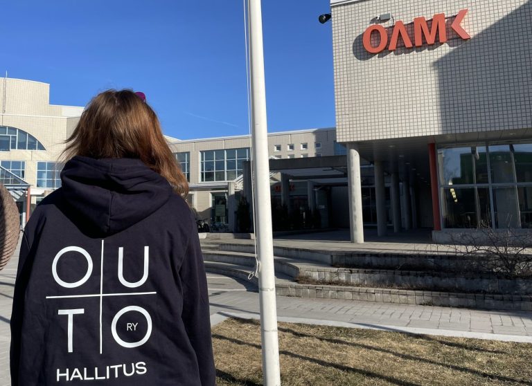 Henkilö selin kameraan OAMK:in pihalla päällään OUTO RY:n hallituksen huppari.