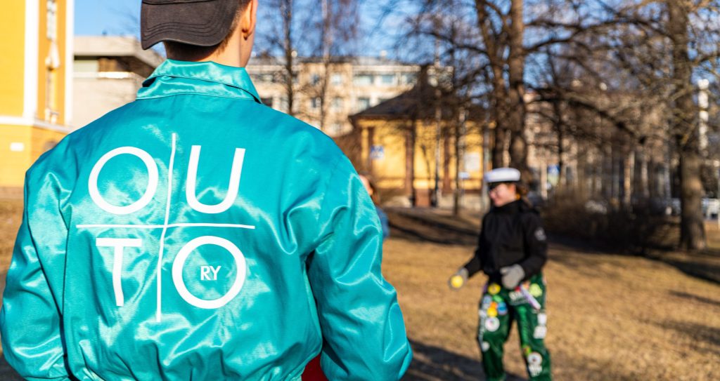 Henkilö turkoosissa Outo ry -haalarissa poseeraa ulkona selin kameraan. Taustalla toinen henkilö vihreissä haalareissa.